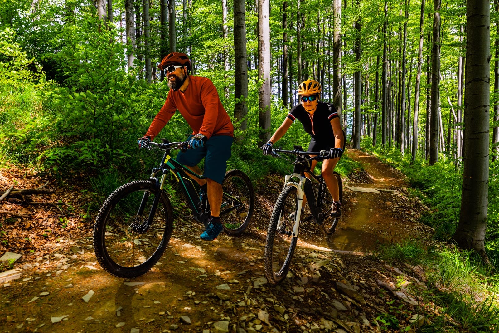 Two people wearing helmets and sunglasses ride mountain bikes on a dirt trail through a sunlit forest.