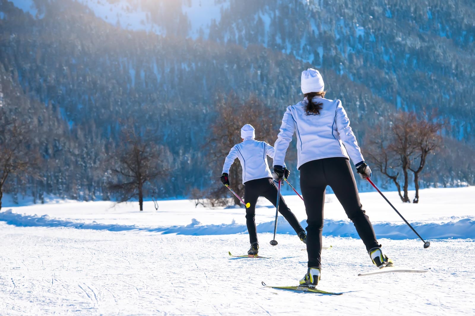 Two people dressed in white jackets and black pants cross-country skiing on a snow-covered trail with mountains and trees in the background.