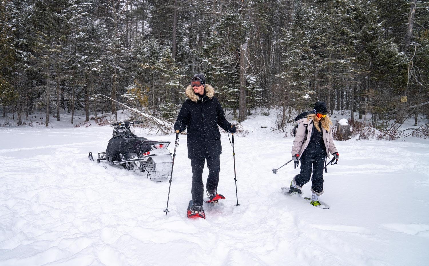 Two people in winter clothing snowshoe in a snowy forest near a snowmobile.